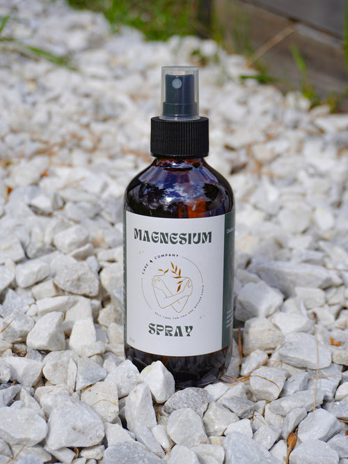 An amber glass bottle of Magnesium Spray. The bottle is placed on a pebble surface outdoors.