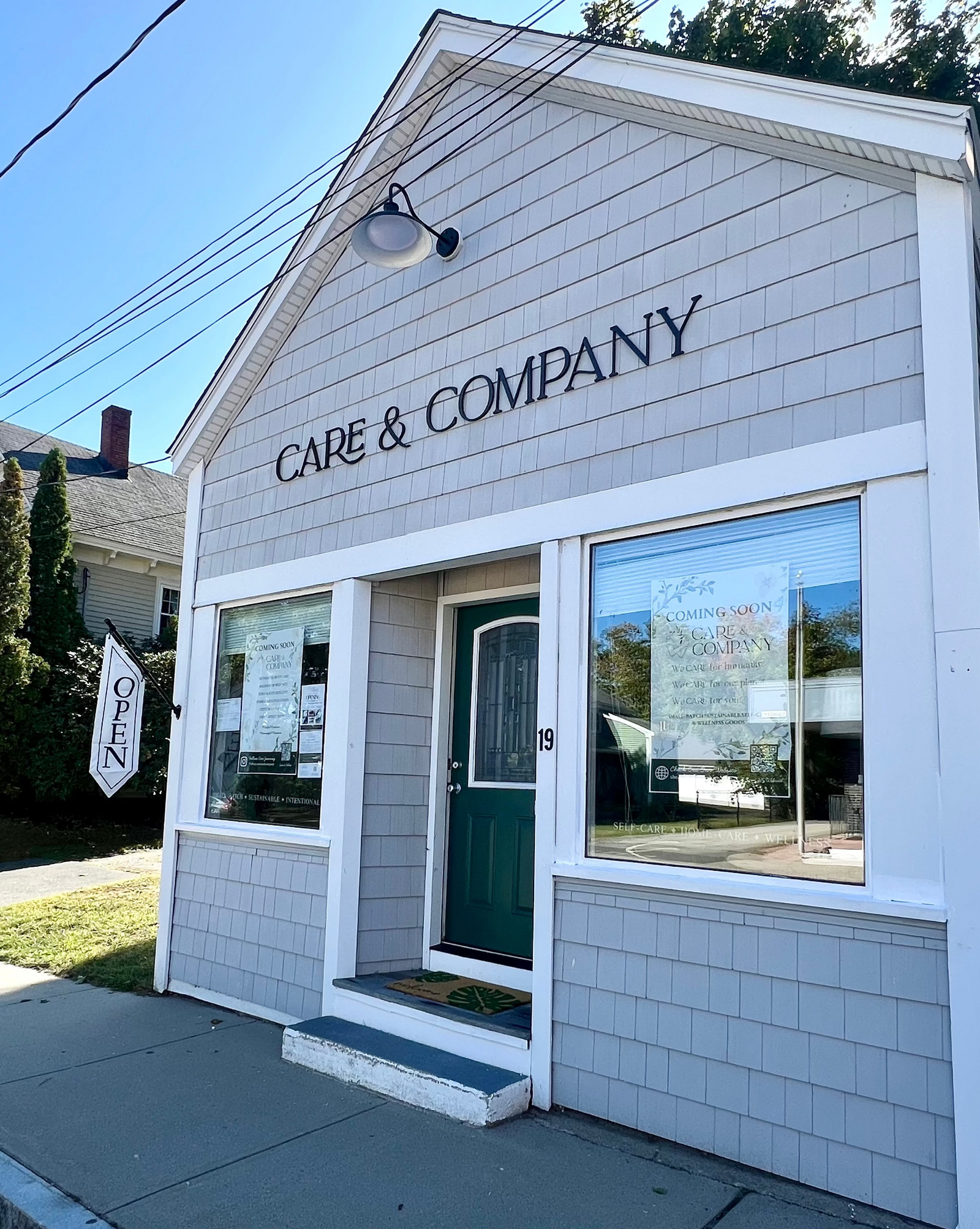 A small white and light gray shingled storefront building with large windows, a dark green front door, and the words 'CARE & COMPANY' above the entrance. An 'OPEN' sign hangs by the window, and the building is photographed outdoors on a sidewalk with trees and sky visible.